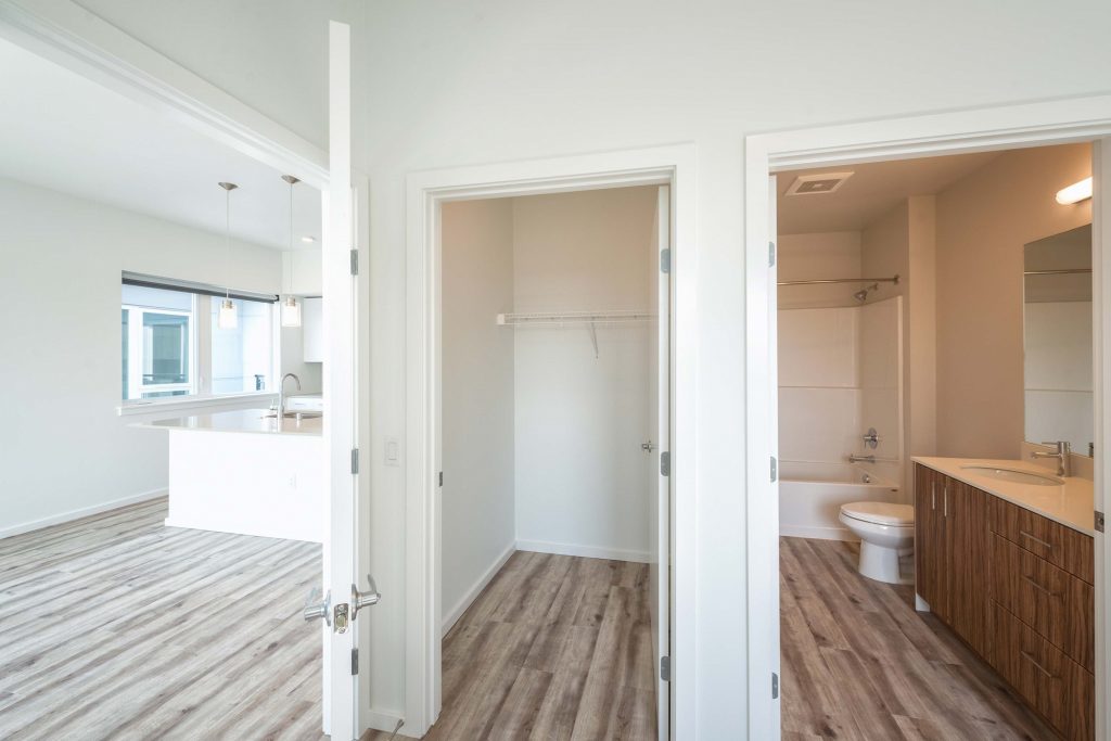 Unfurnished hallway with open closet, adjacent open door to bathroom, and adjacent open doorway to living room and kitchen